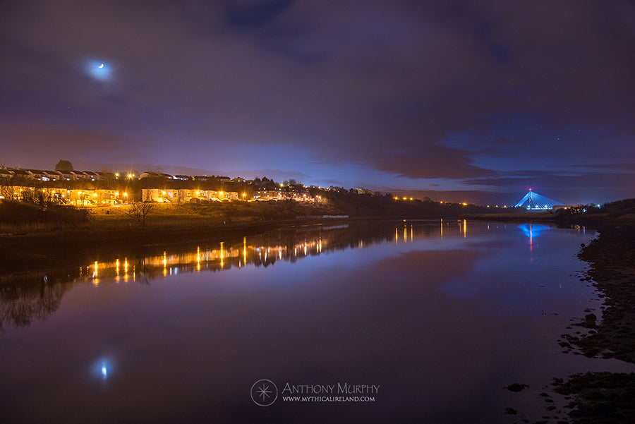 Blue hour along the Boyne. The crescent moon is reflected in the water in this image showing River Court and the Boyne Valley (Cable) Bridge.The image was taken along the public pathway that hugs the river Boyne at Mell, along the northern fringes of the river. The Mary McAleese Boyne Valley Bridge (known since it was built as the Boyne Cable Bridge) can be seen in the distance.