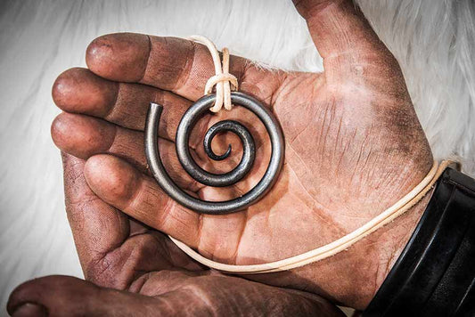 This image shows a hand forged spiral pendant on a leather necklace, held in the working hands of a smith.