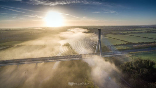 An aerial image of the Boyne Cable Bridge which carries the M1 motorway across the Boyne river west of Drogheda, on a foggy morning at sunrise. The morning mist and fog tends to form close to the river, and this provides opportunities for some gorgeous photography. In this image, it is as if the river itself has become a floating stream of fog, running beneath the great span of the bridge.